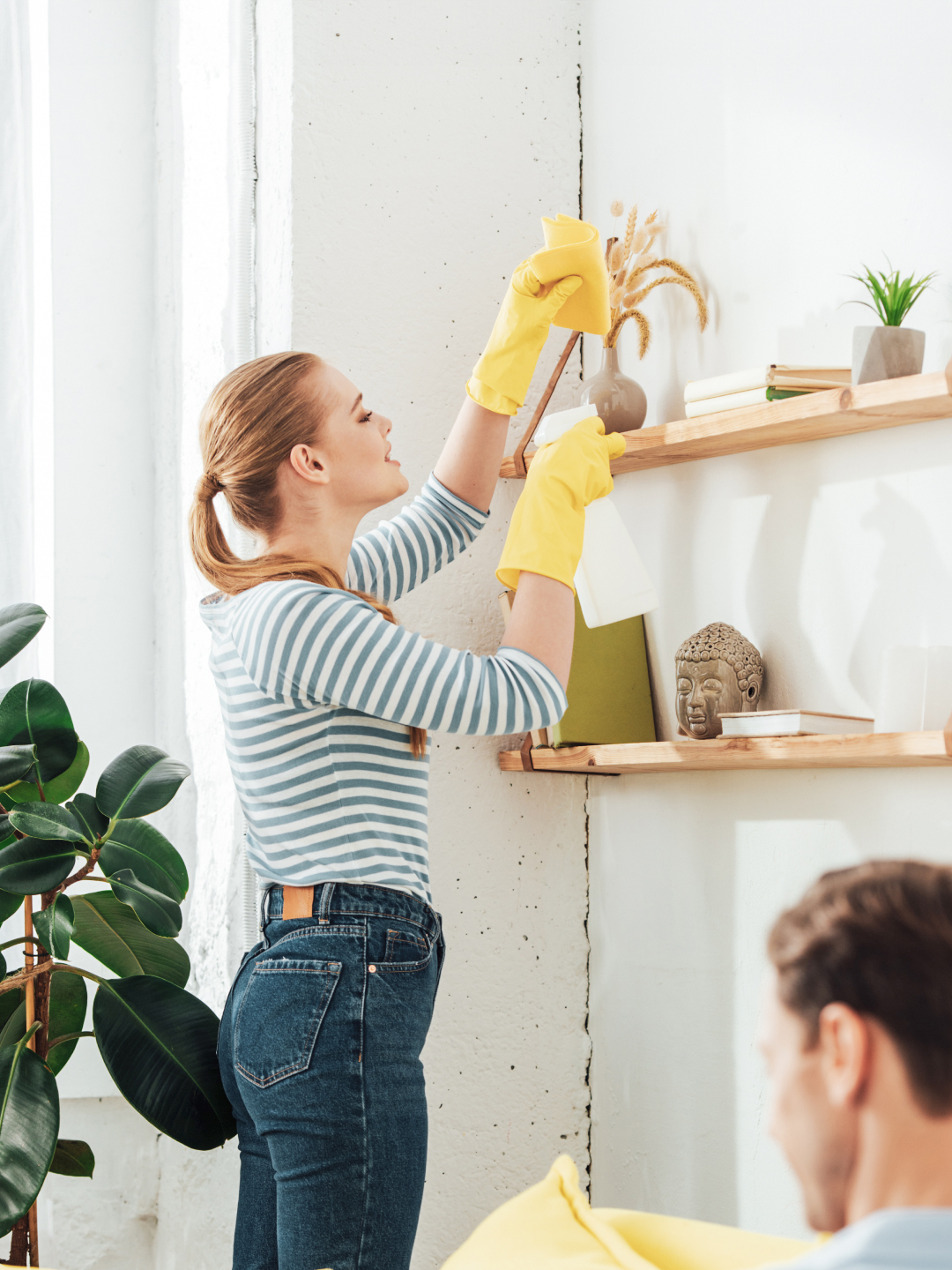 A woman wearing yellow rubber gloves is cleaning a shelf in a bright, modern living space. She is arranging decorative items and plants on the shelf, while a man sits casually on a couch in the background.