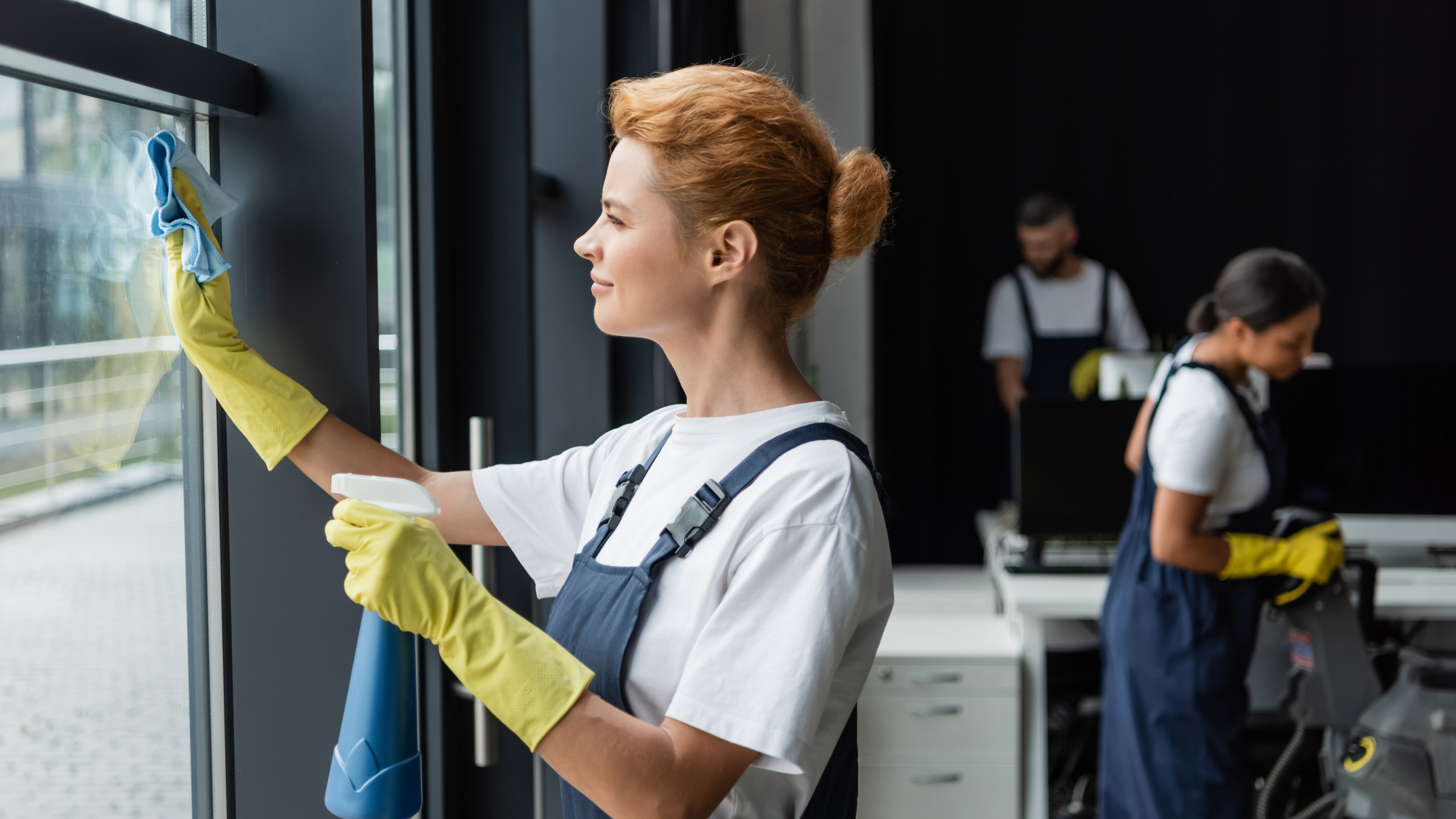 A woman in a white shirt and yellow gloves is cleaning a window with a cloth, while two people in the background are tidying up an office space. One is using a vacuum cleaner, and the other is working at a desk.