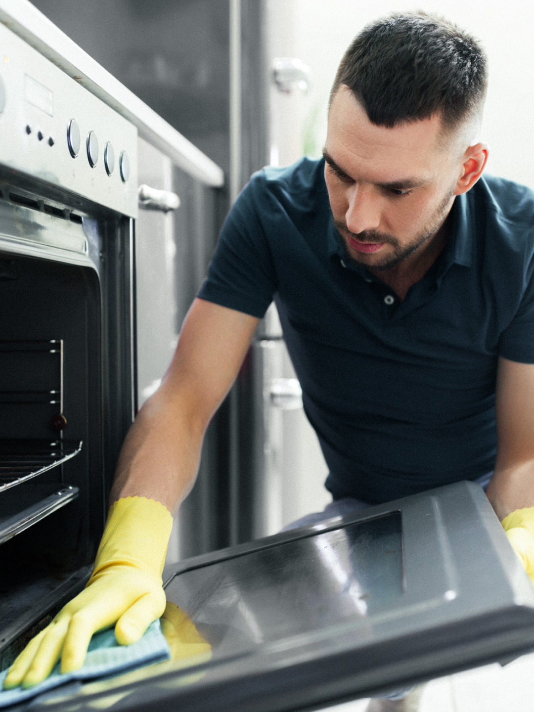 A man in a blue polo shirt and yellow gloves is cleaning the interior of an oven. He is focused on wiping the glass door of the oven with a cloth while kneeling on the floor beside it.